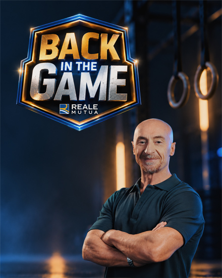 Primo piano di Jury Chechi sorridente con a fianco il logo del format 'Back in the game' e il logo di Reale Mutua, sullo sfondo una palestra e l'attrezzo con i cerchi, specialit&agrave; sportiva del grande campione