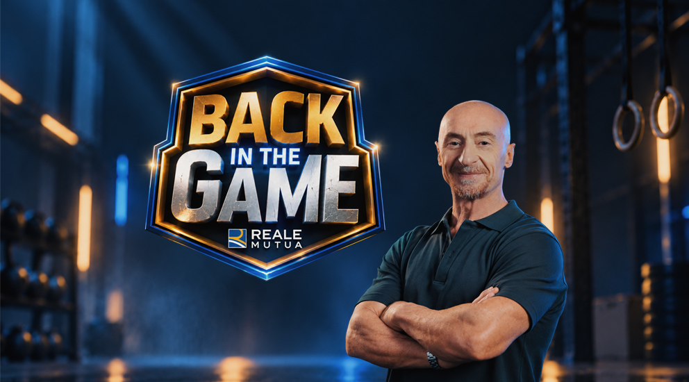 Primo piano di Jury Chechi sorridente con a fianco il logo del format 'Back in the game' e il logo di Reale Mutua, sullo sfondo una palestra e l'attrezzo con i cerchi, specialità sportiva del grande campione