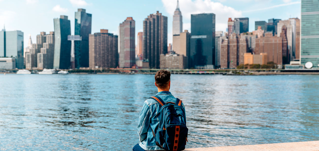 Ragazzo visto di spalle che indossa uno zaino, seduto su un muretto davanti al fiume e sullo sfondo lo skyline di New York