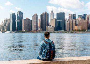Ragazzo visto di spalle che indossa uno zaino, seduto su un muretto davanti al fiume e sullo sfondo lo skyline di New York
